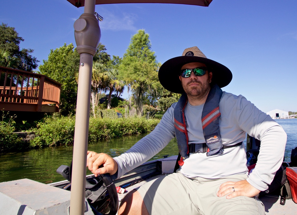 Man wearing a wide-brimmed hat, sunglasses, and a backpack sitting on a boat near water with trees in background.