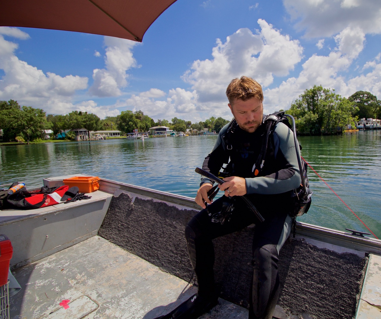 Man sitting on boat with water and trees in background, holding a device, under a red canopy.
