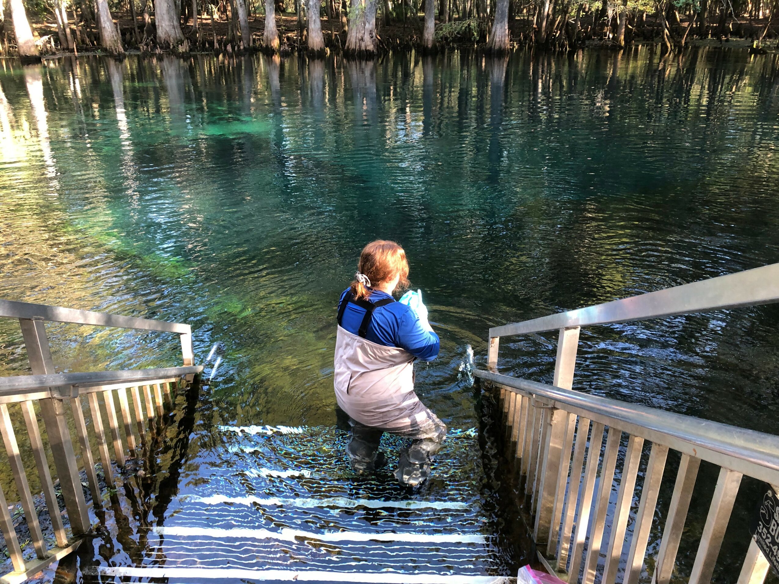 Child wading in water near stairs with metal railings, trees in background, sunny day.