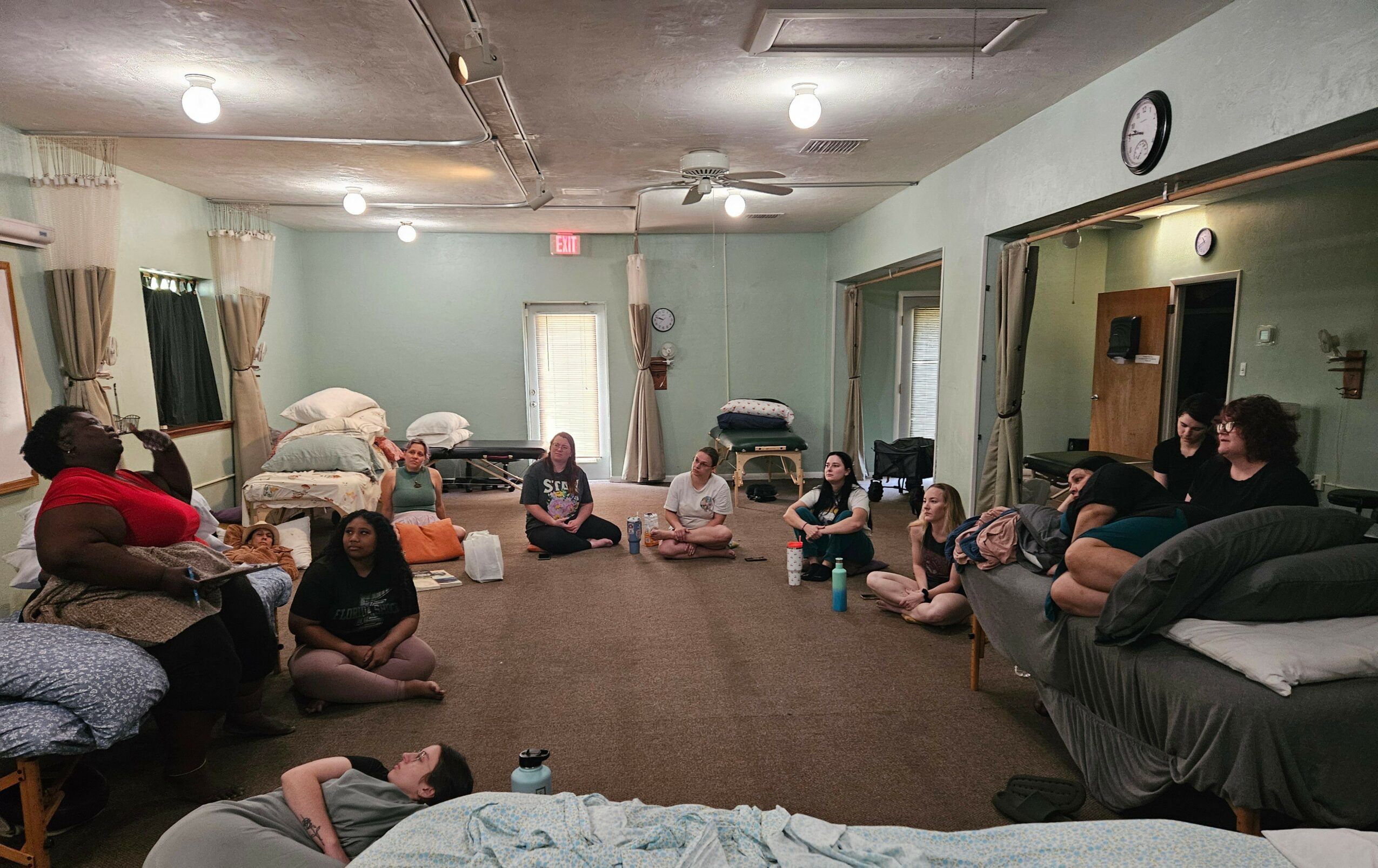 Group of people sitting on the floor in a large room, some with pillows, facing each other in a circle.