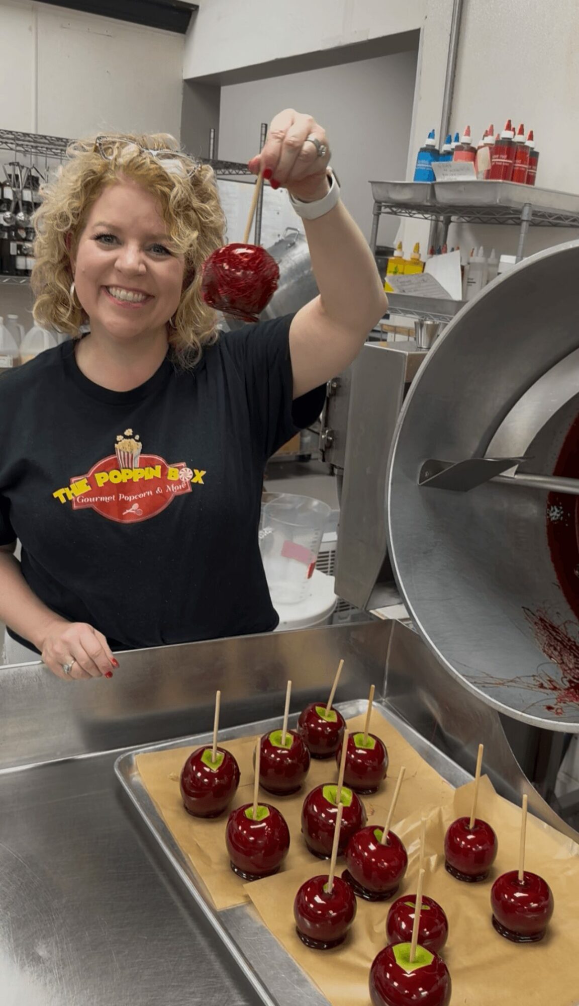 Woman with curly blonde hair smiling, holding a cherry on a stick in a kitchen with cherry desserts on a tray.