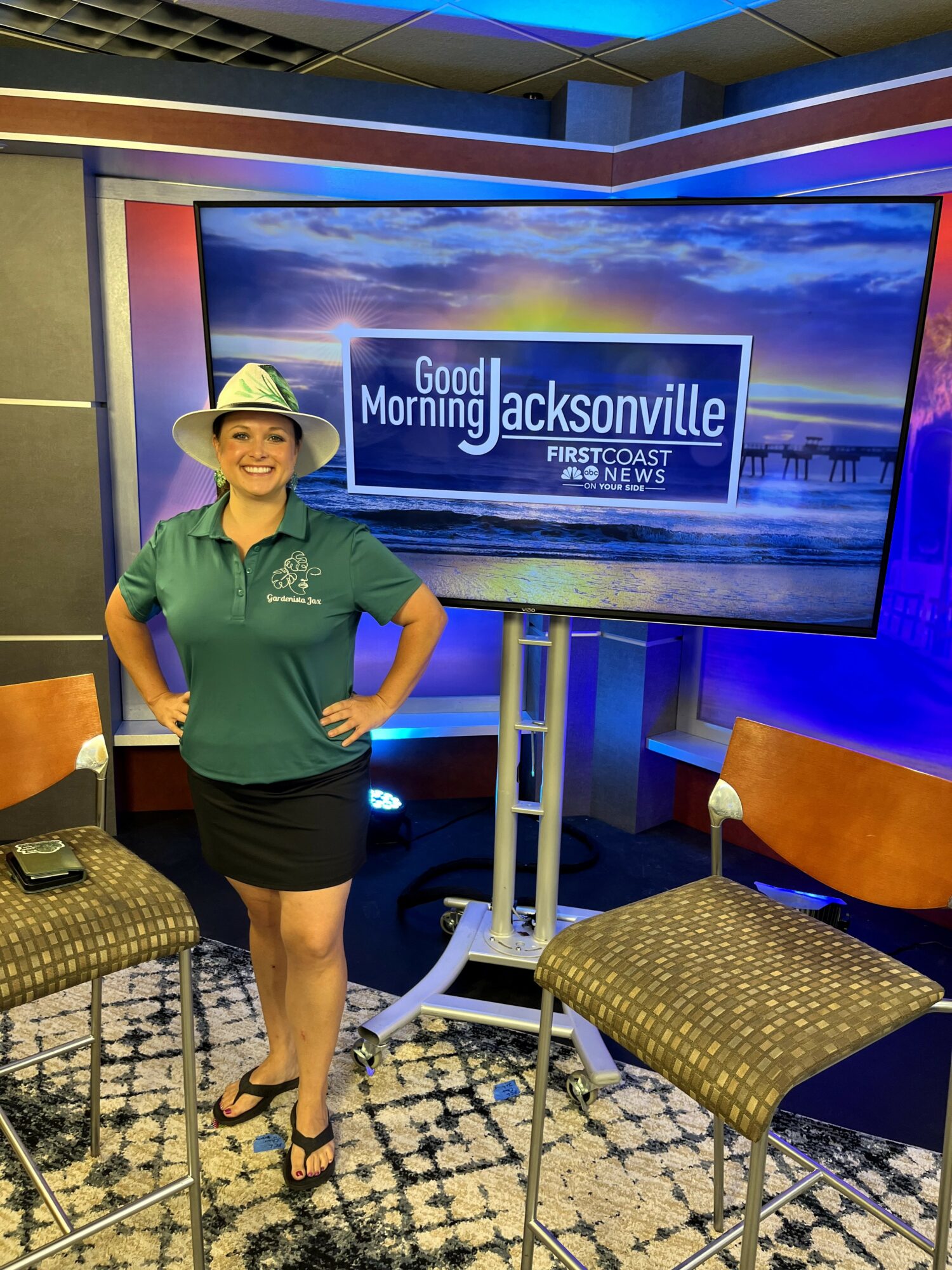 Woman in green shirt and hat standing next to a TV screen displaying 'Good Morning Jacksonville' news program.