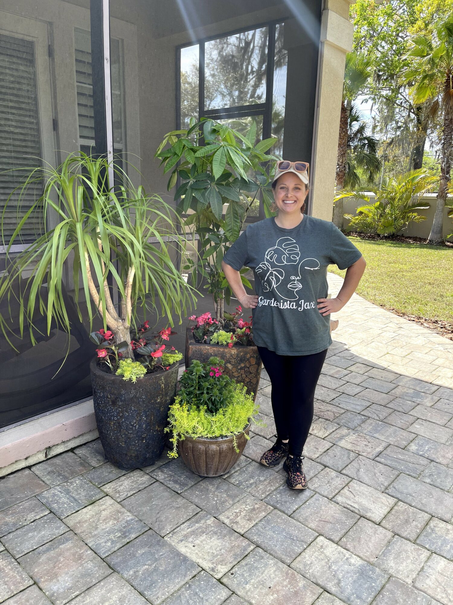 Girl standing outdoors next to potted plants on a paved area, smiling, wearing a cap and dark clothing.