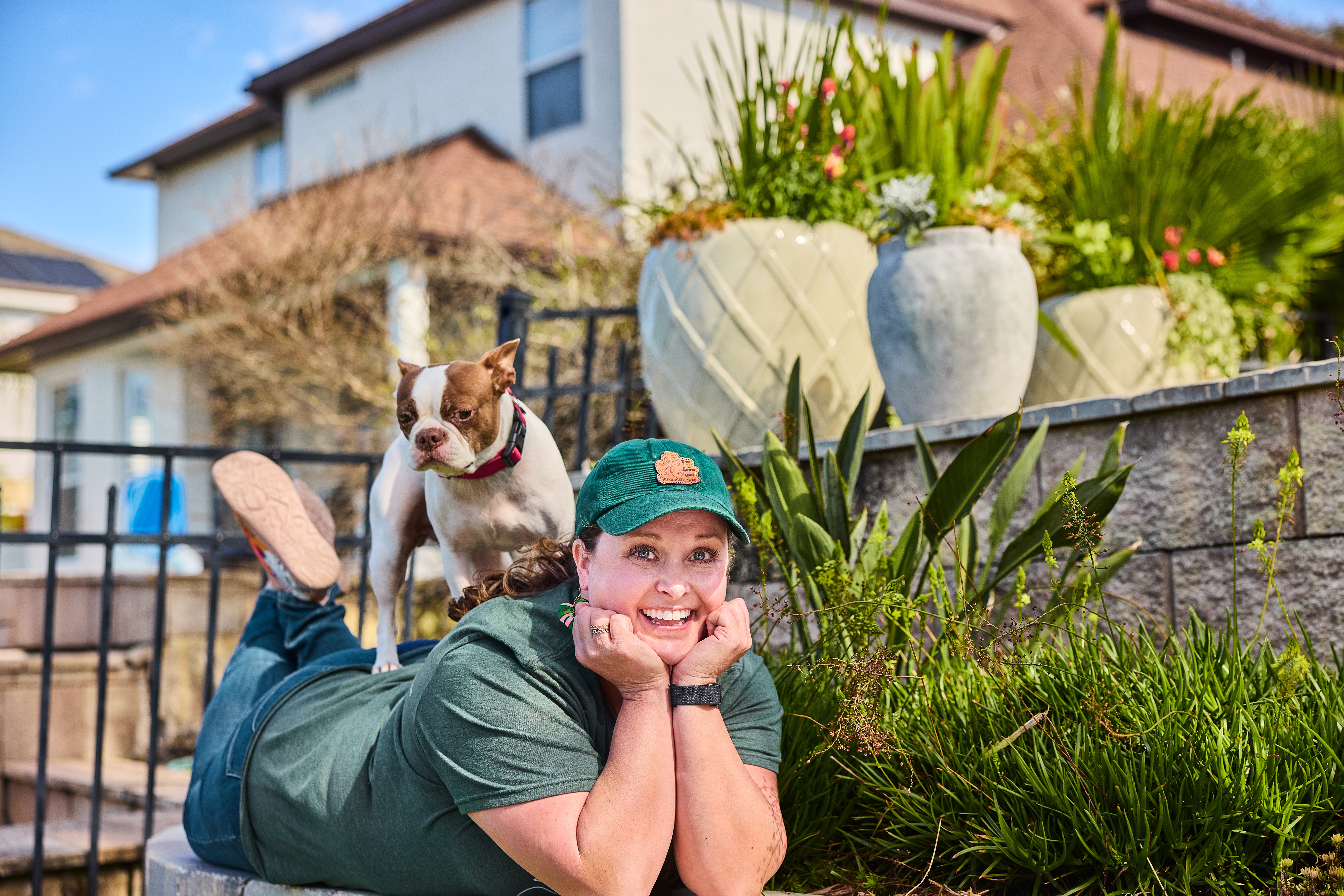 Woman lying on grass with a small dog on her back, smiling, in a garden with potted plants and a house in the background.