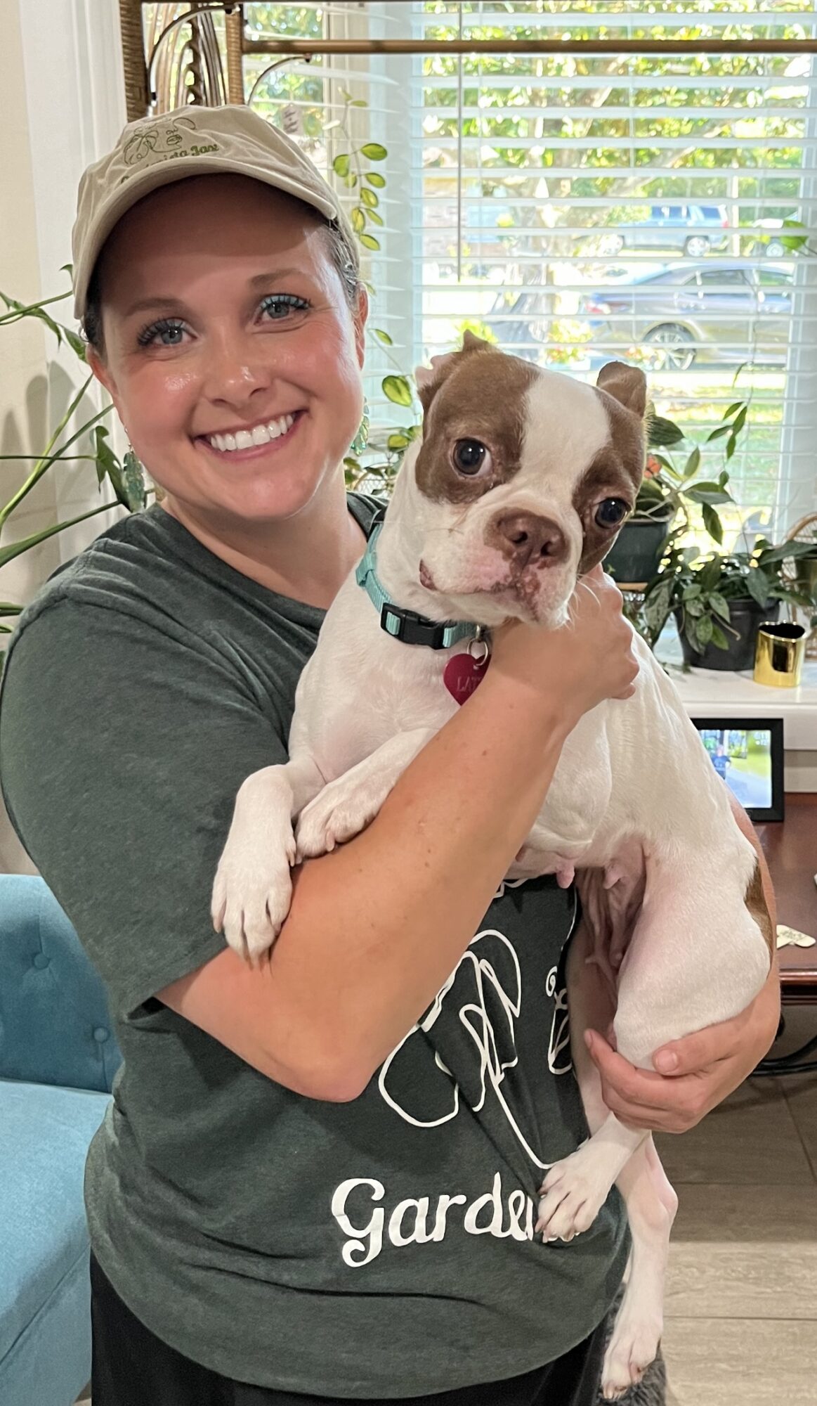 Woman smiling holding a small dog indoors near a window with plants.