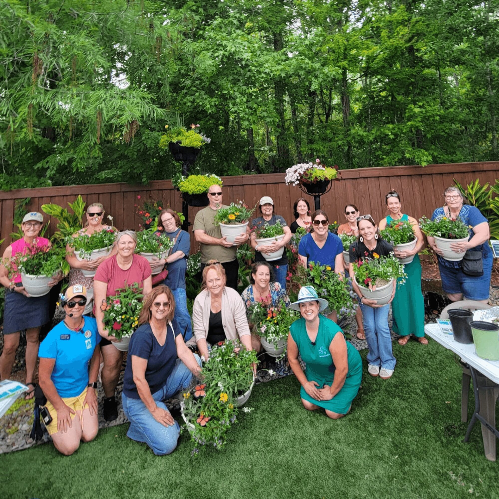 Group of people outdoors holding potted plants and flowers, standing and sitting on grass with a wooden fence and trees behind.