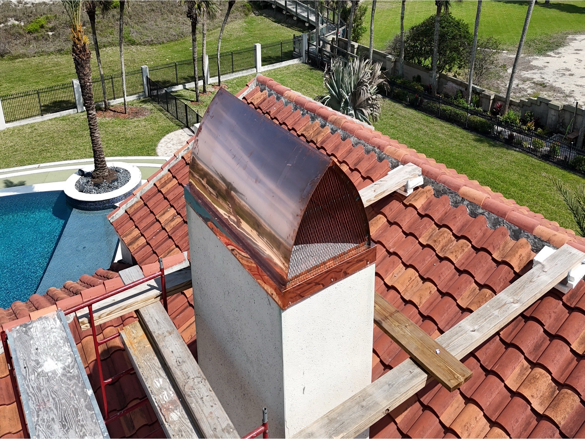 A chimney with a curved metal cap on a red-tiled roof, surrounded by a garden and pool area.