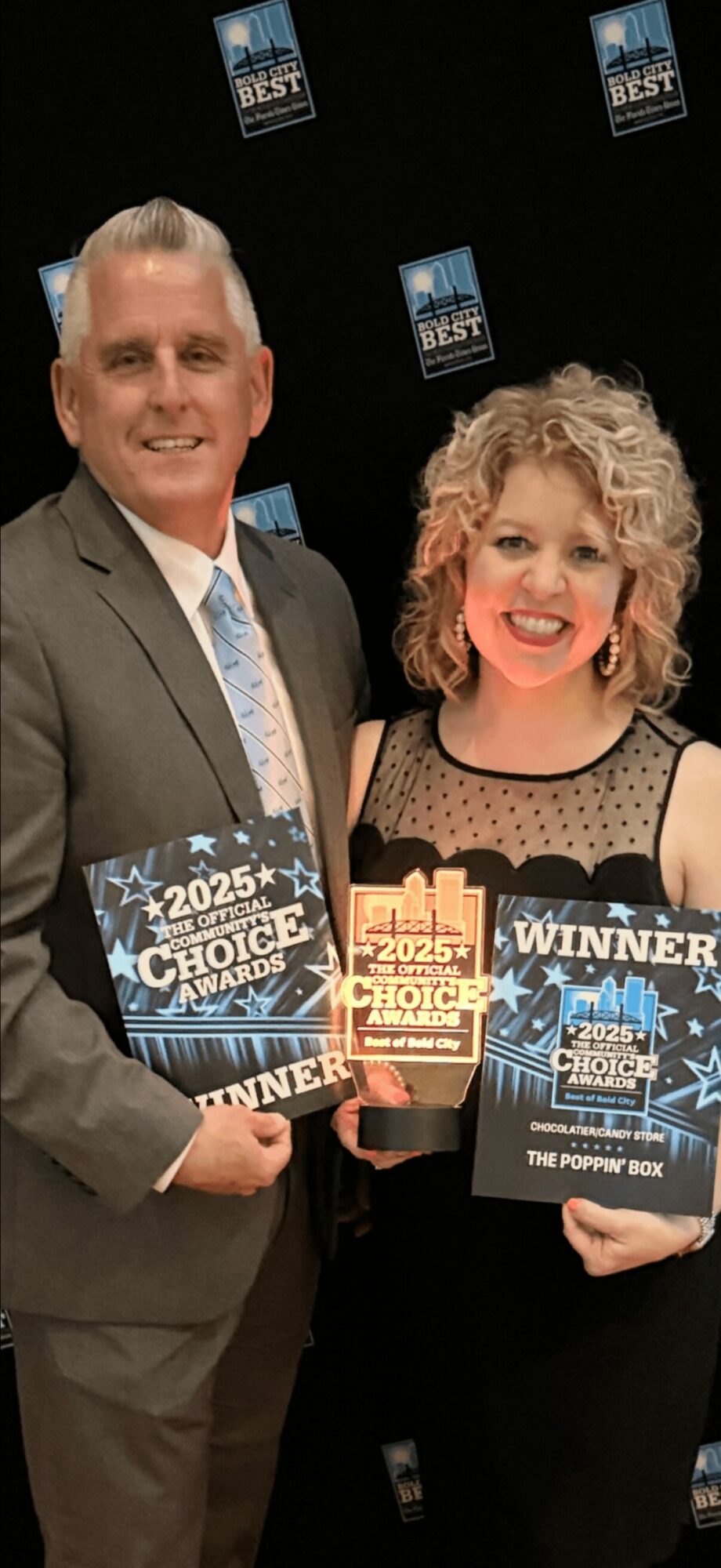 Two people holding awards and certificates, smiling, at an event with a black background and event logos.