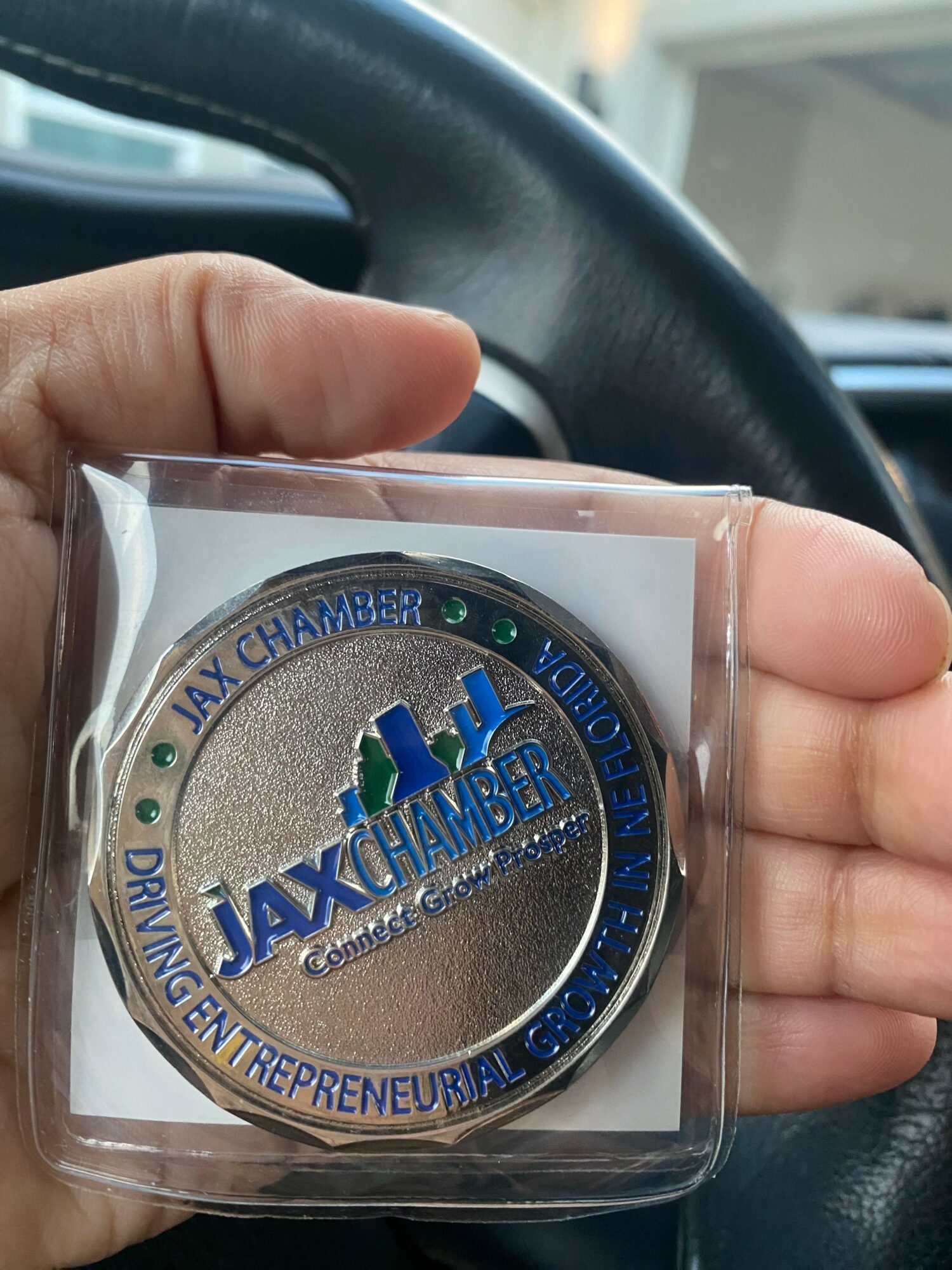 Hand holding a circular badge with blue and silver design, text, and city skyline, inside a plastic case.