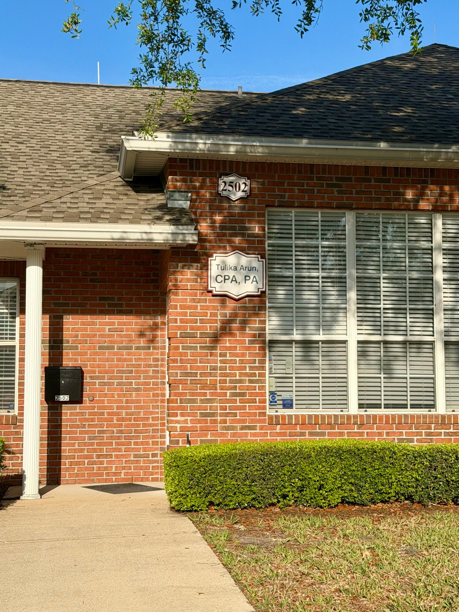 Brick house with large window, hedge, and address signs, under a blue sky with trees.