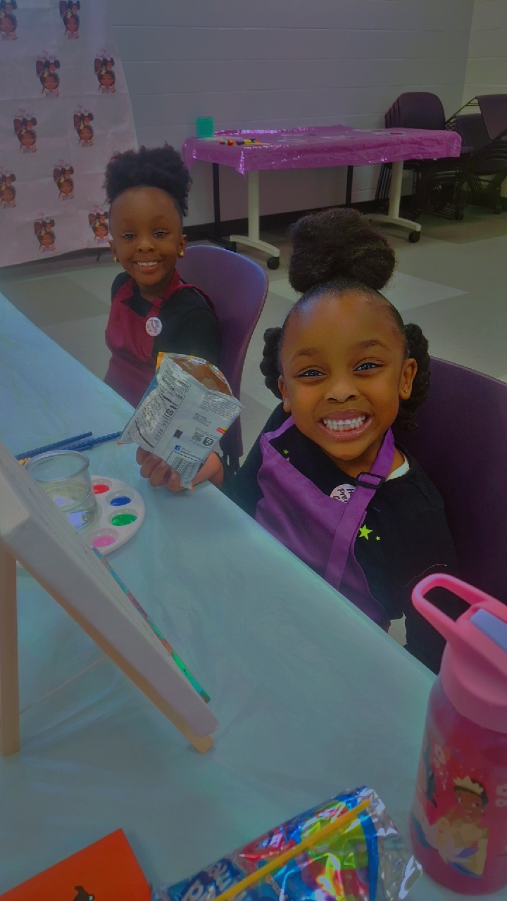 Two children smiling at a table with art supplies, one holding a decorated object, in a room with chairs and tables.