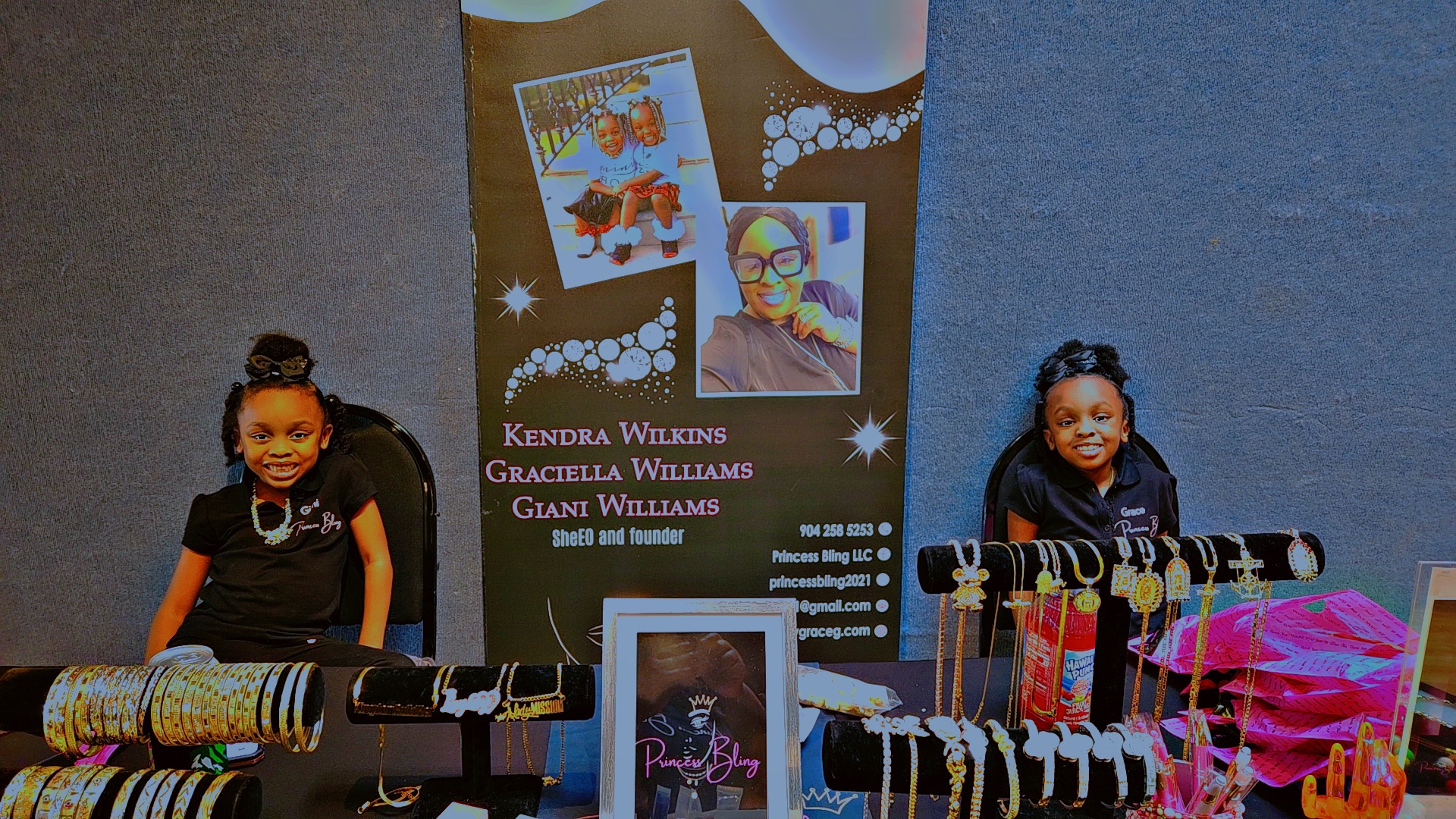 Two women sitting at a table with colorful jewelry displays and a promotional banner behind them.