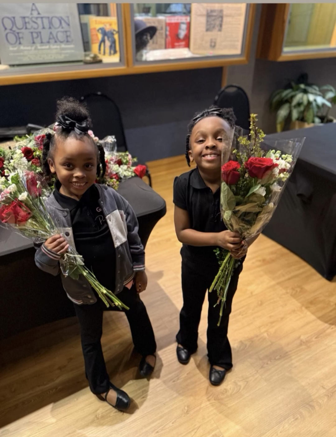Two young girls holding bouquets of red and white flowers, smiling indoors with books and plants in the background.