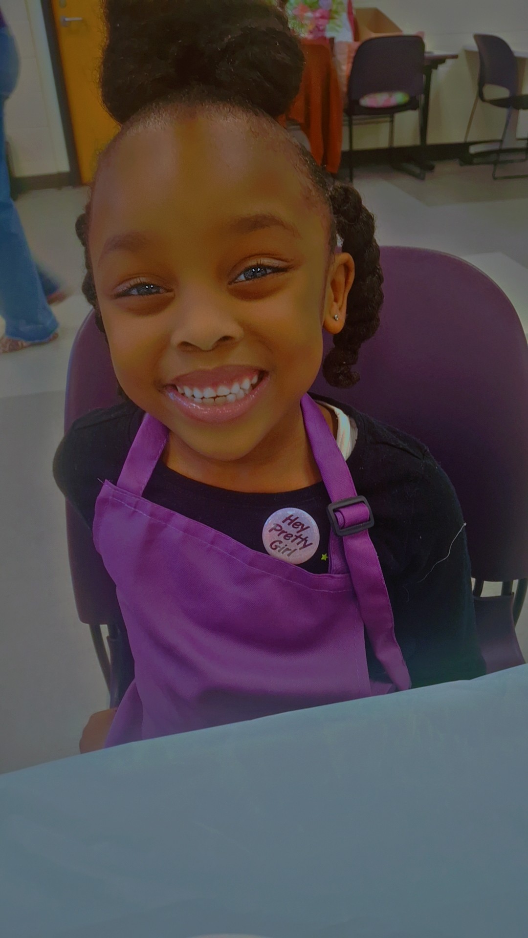 Young girl smiling, wearing a purple apron, sitting at a table in a room with chairs and people in the background.