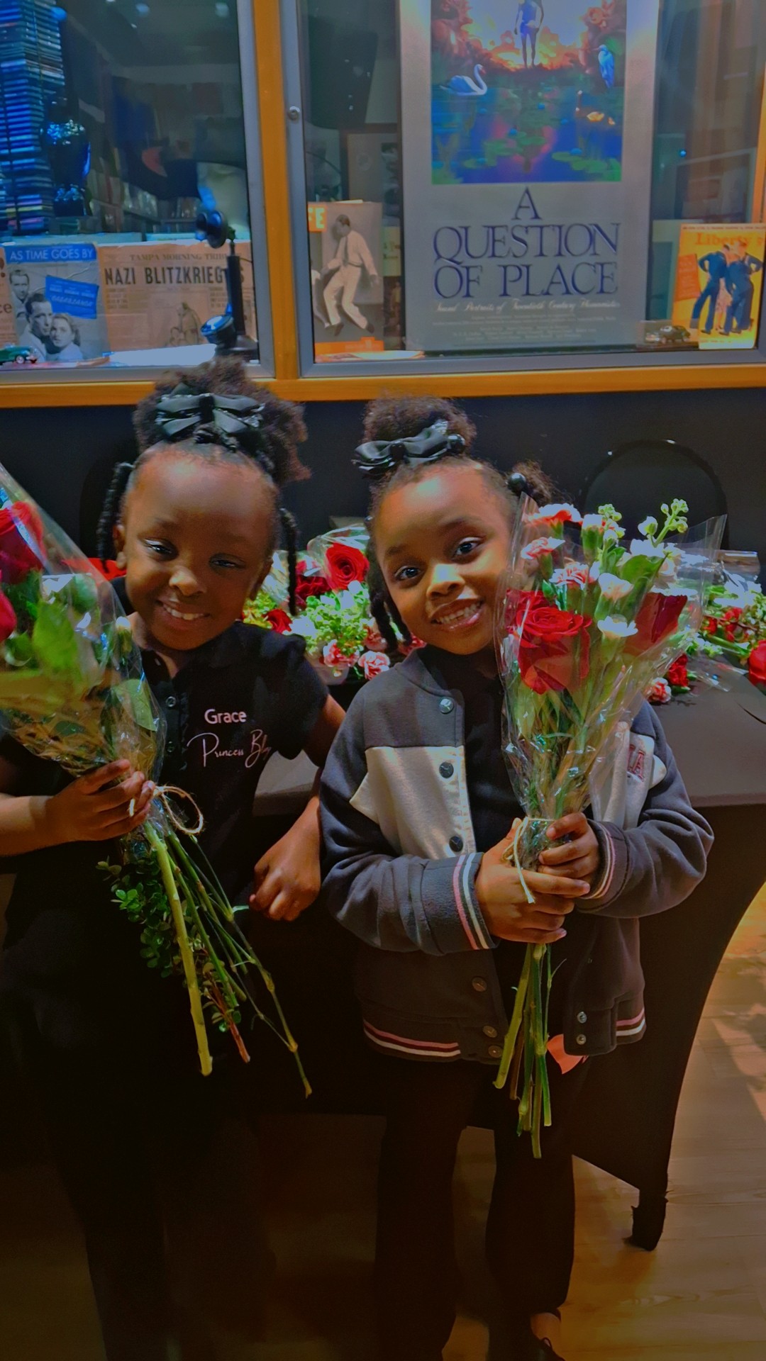 Two children holding bouquets of flowers, smiling, standing in front of a display with books and flowers.