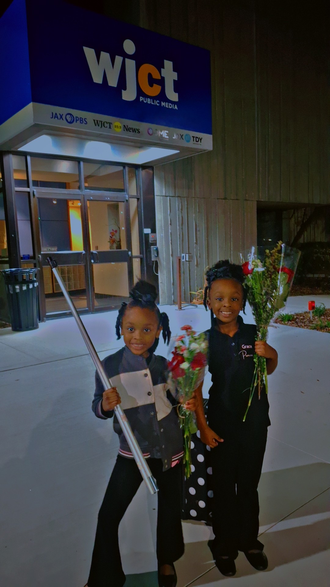 Two children stand outside the entrance of a building with a sign reading 'wjct', holding flowers and a stick.