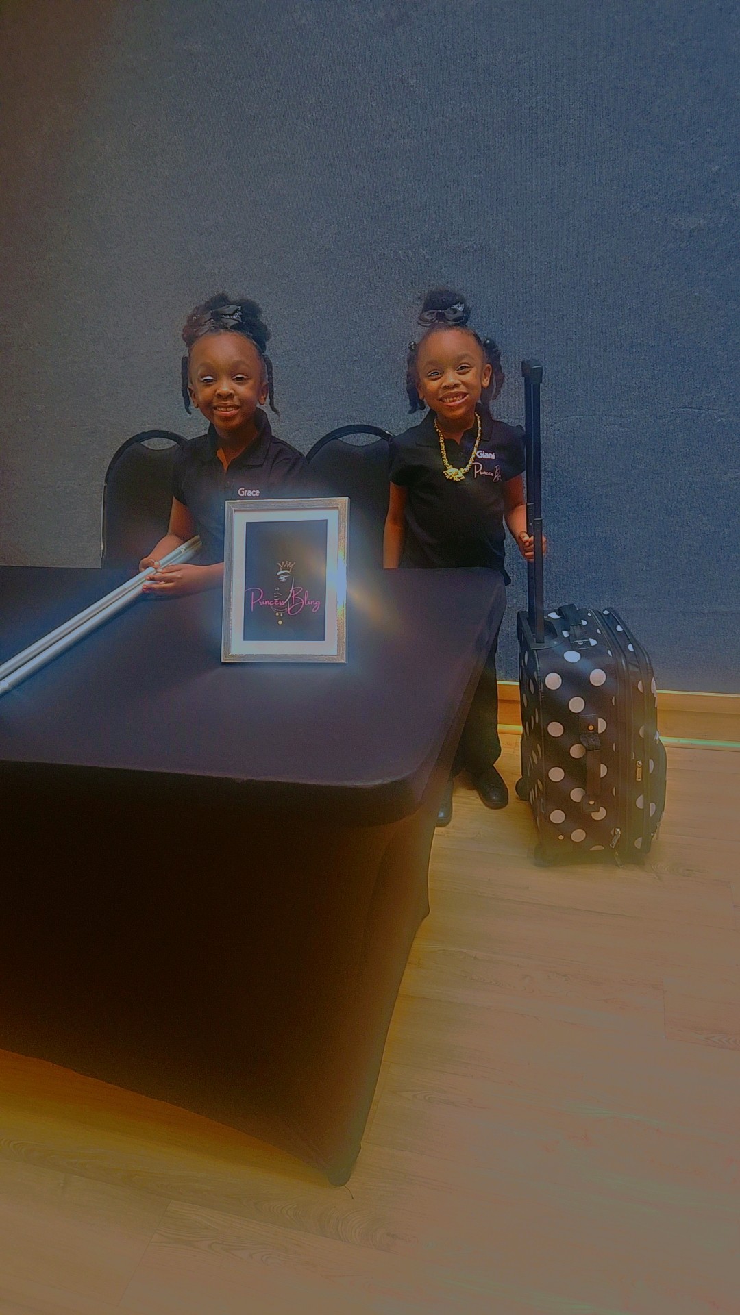 Two children sit at a table with a framed picture, a suitcase, and a blue wall in the background.