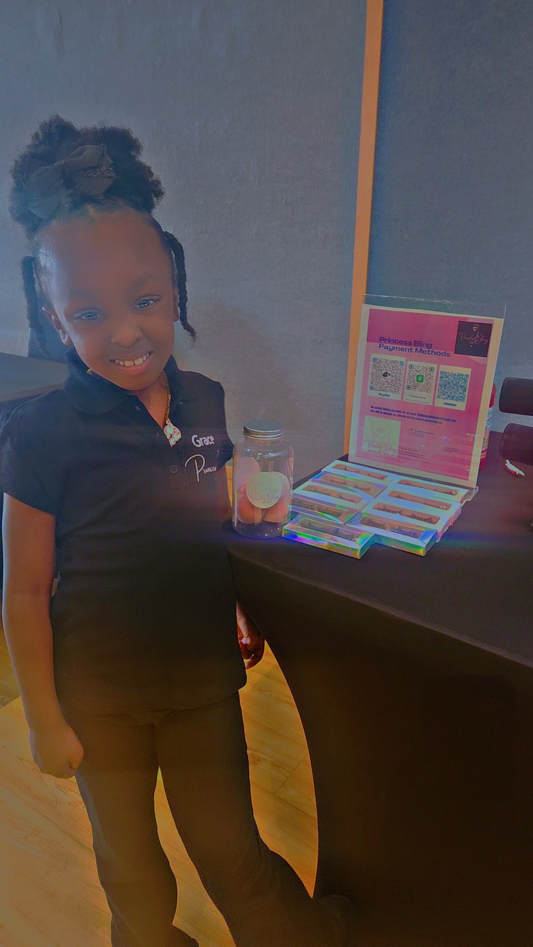 Young girl with braided hair holding a drink, smiling, standing next to a table with a colorful box and laptop.