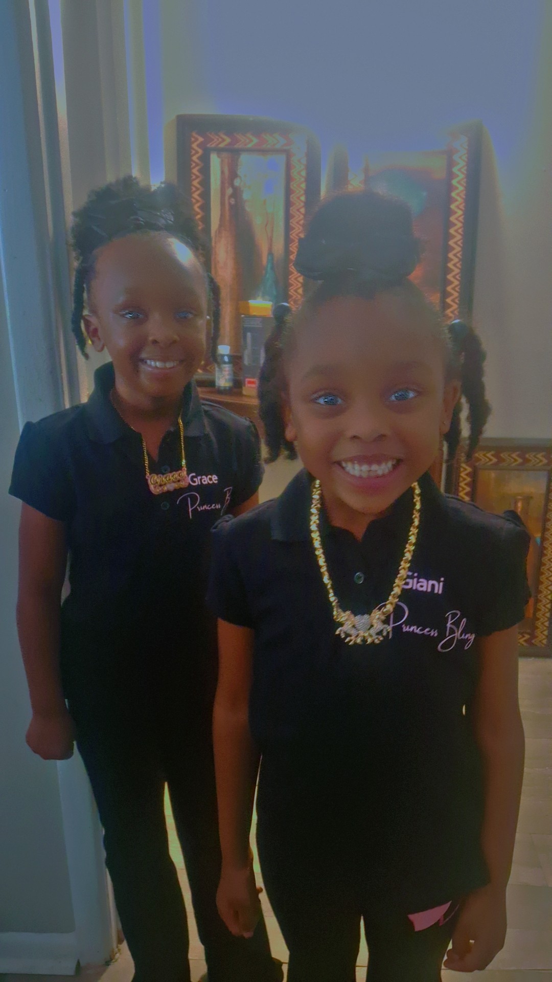 Two smiling children with braided hair and dark uniforms standing indoors, with chairs and a window in the background.
