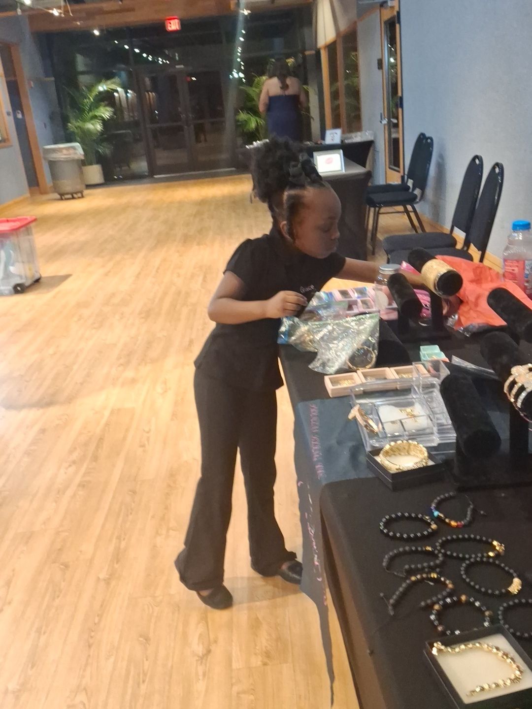 Child with curly hair browsing jewelry display in store, wooden floor, glass windows, and chairs in background.