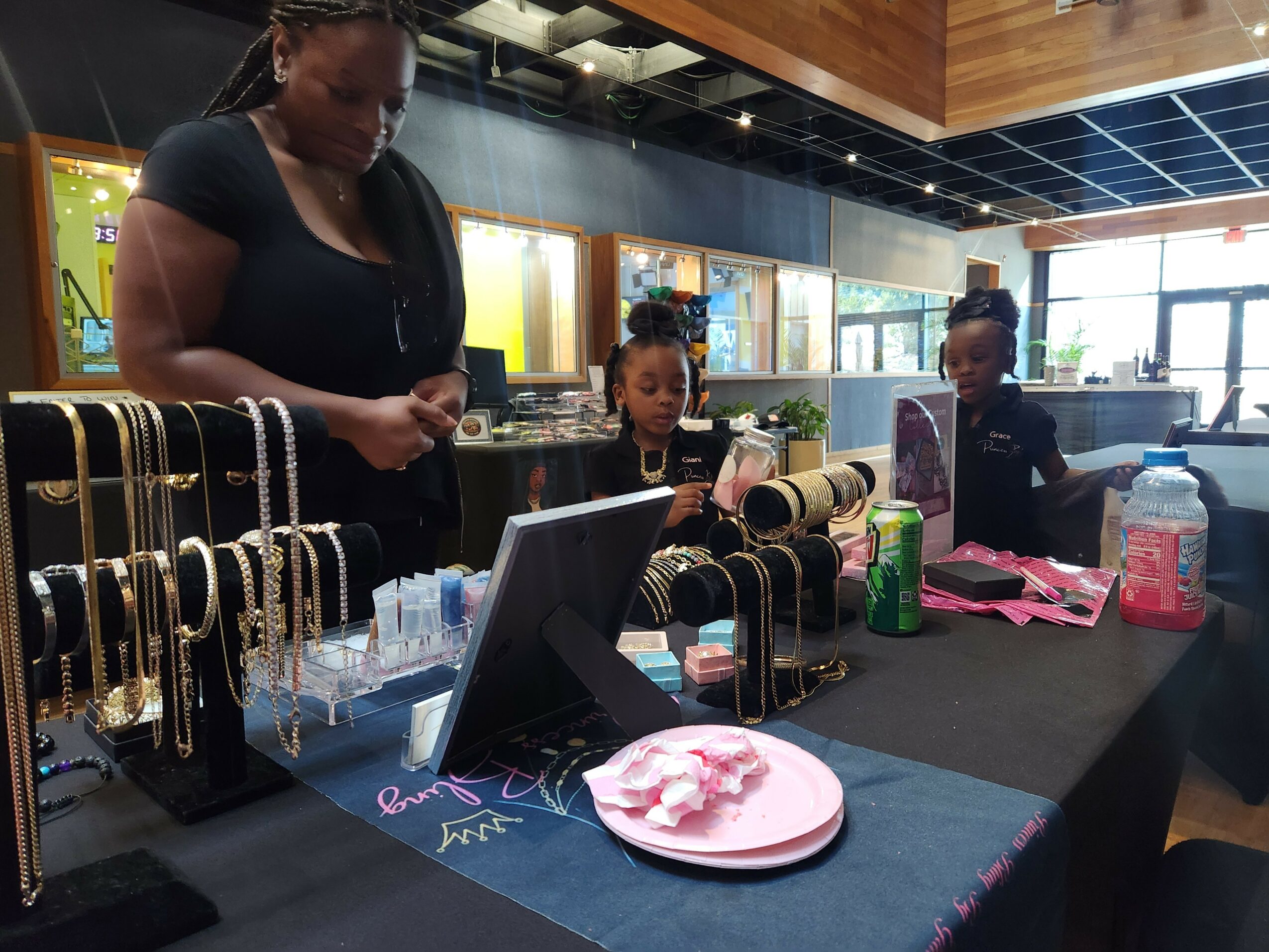 Woman and three children browsing jewelry display in store, with necklaces and accessories on table.