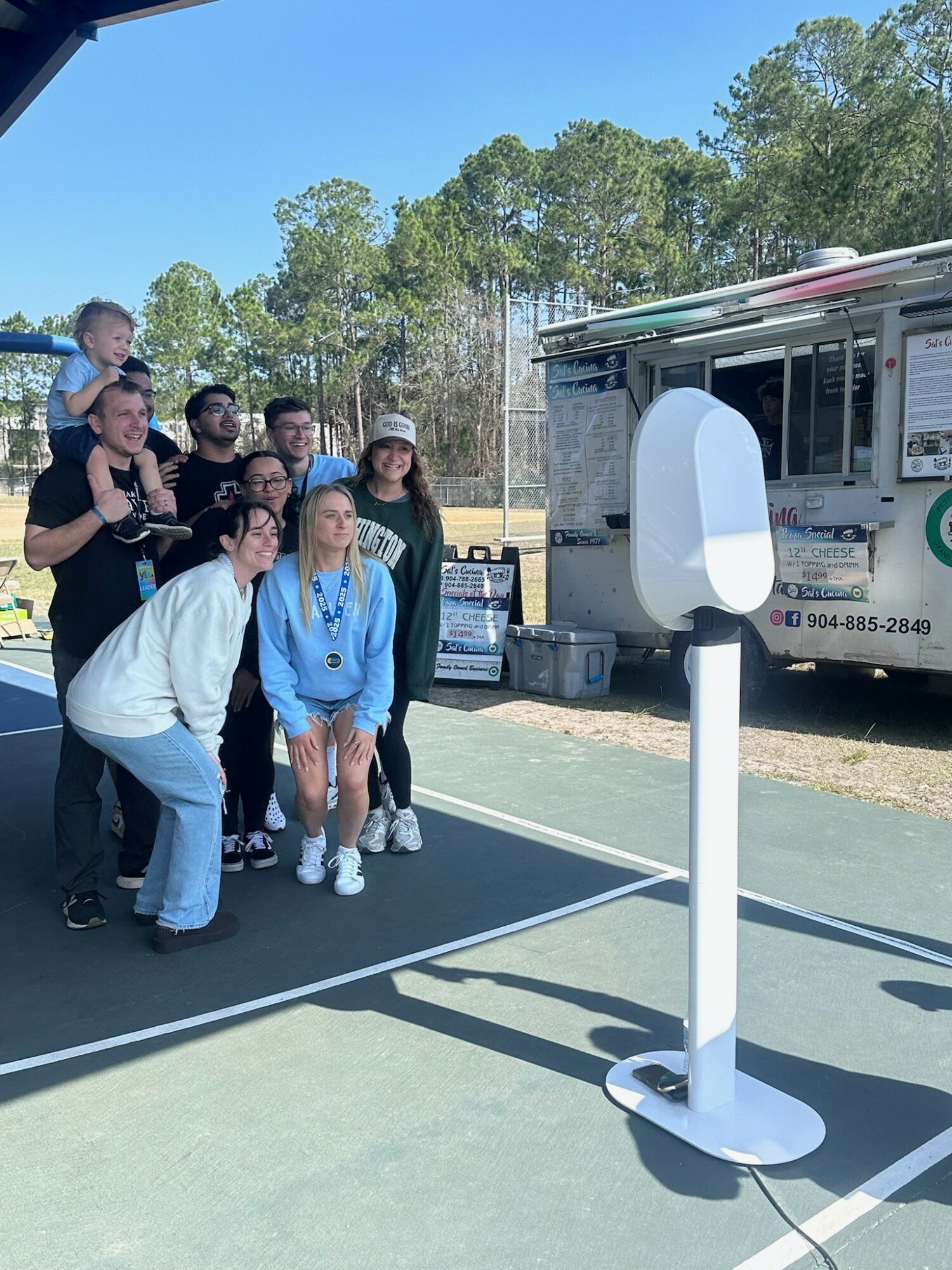 Group of people posing for a photo in front of a food truck outdoors with trees in the background.