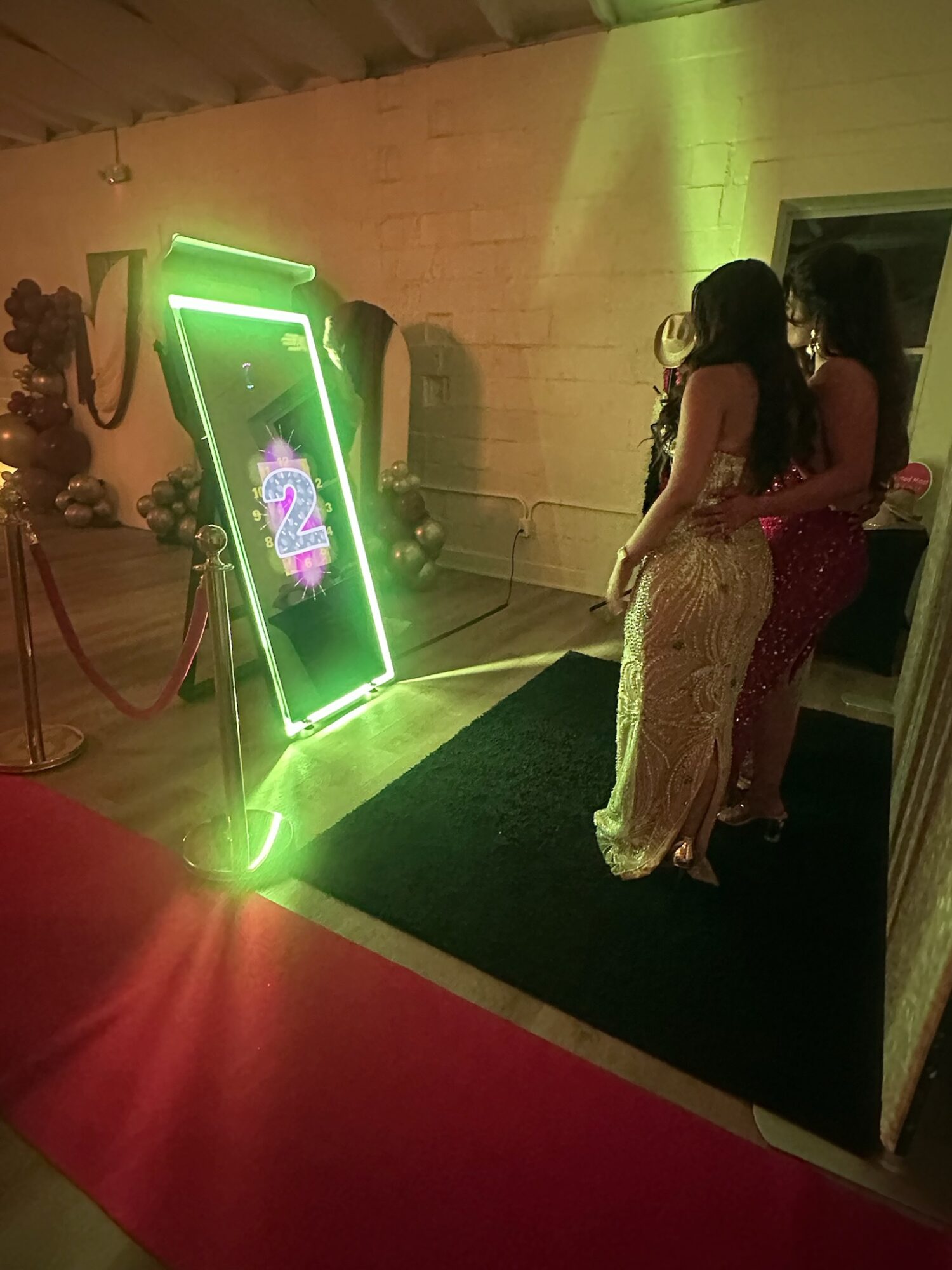 Two women in elegant dresses stand on a black platform with a green neon sign nearby, in a dimly lit room.