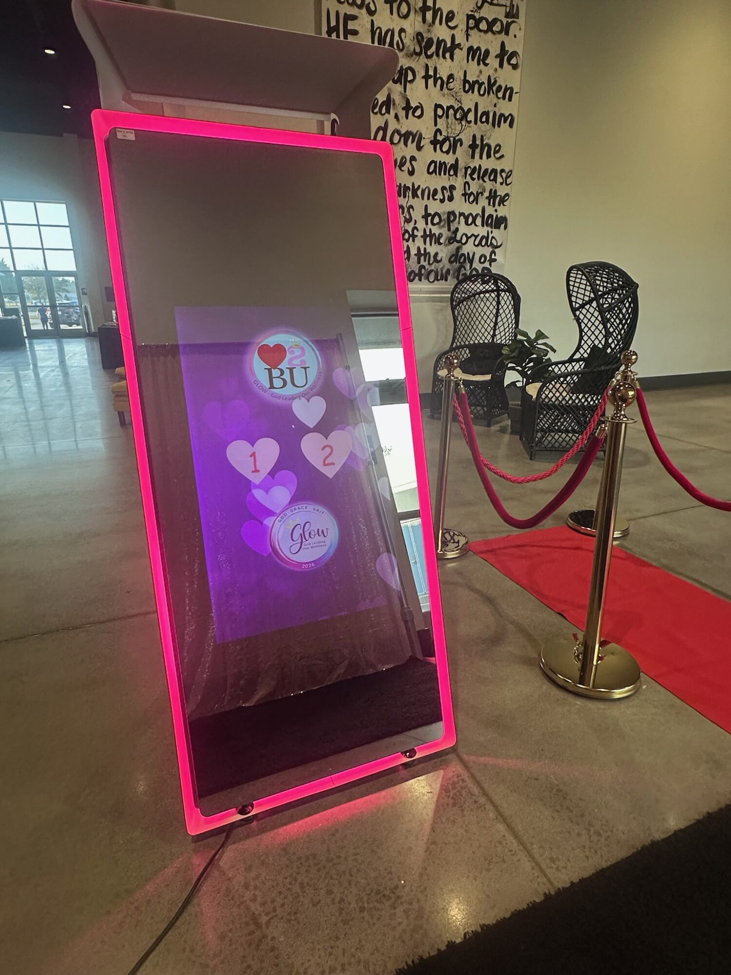 Pink-framed mirror with digital display showing hearts and text, placed on a tiled floor near chairs and a red carpet.