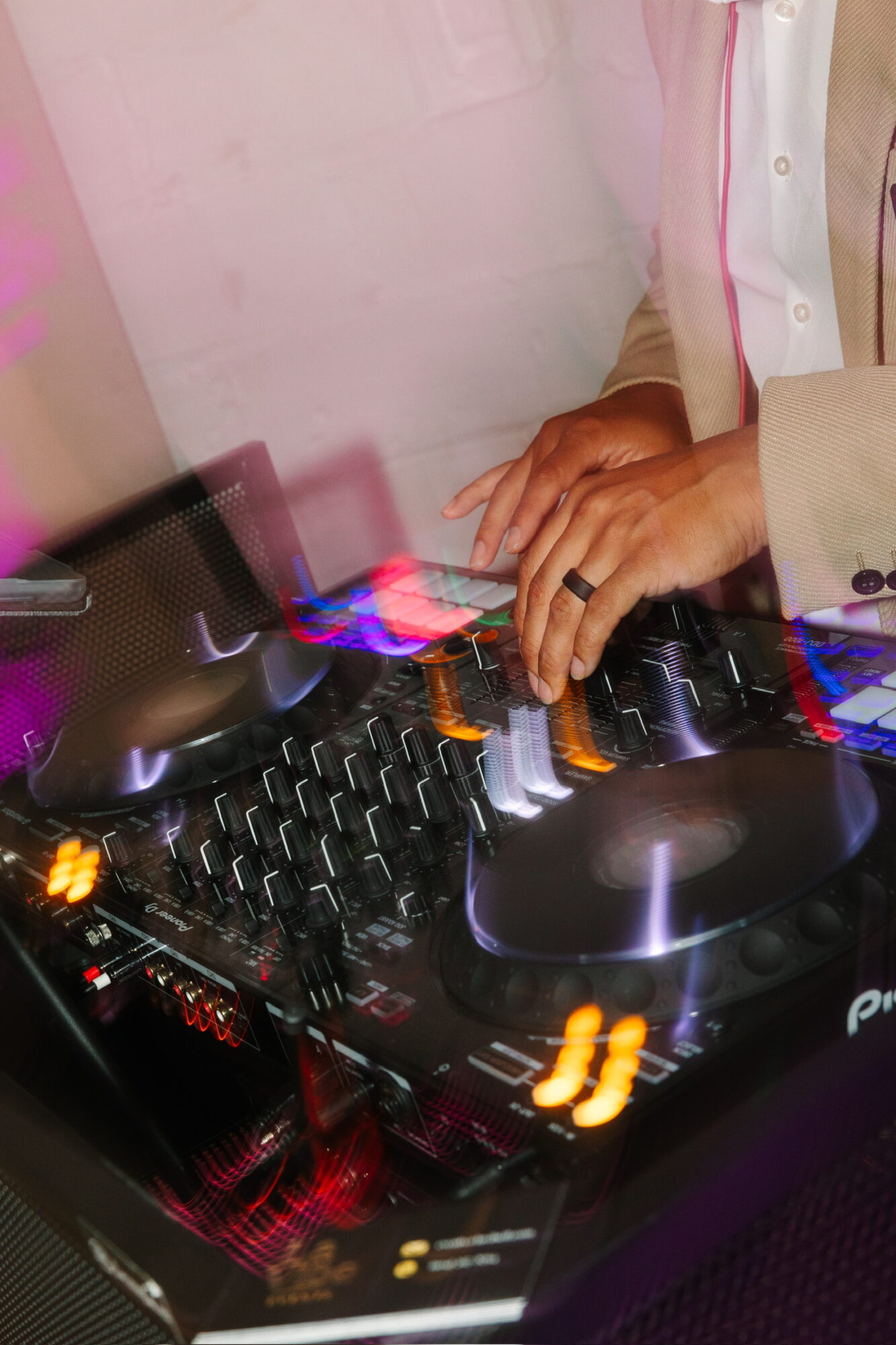 Person's hands operating DJ equipment with colorful lights and controls, wearing a beige jacket with pink accents.
