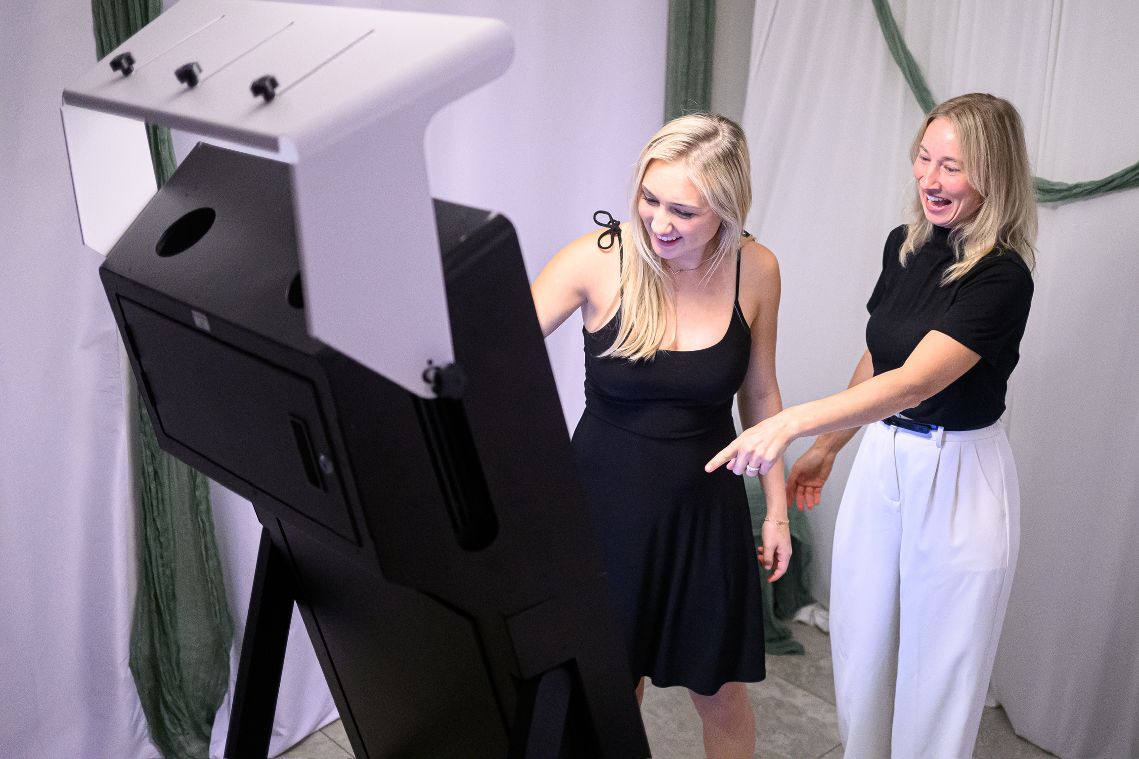 Two women smiling and pointing at a large black photo booth or kiosk in a room with white curtains and green decorations.