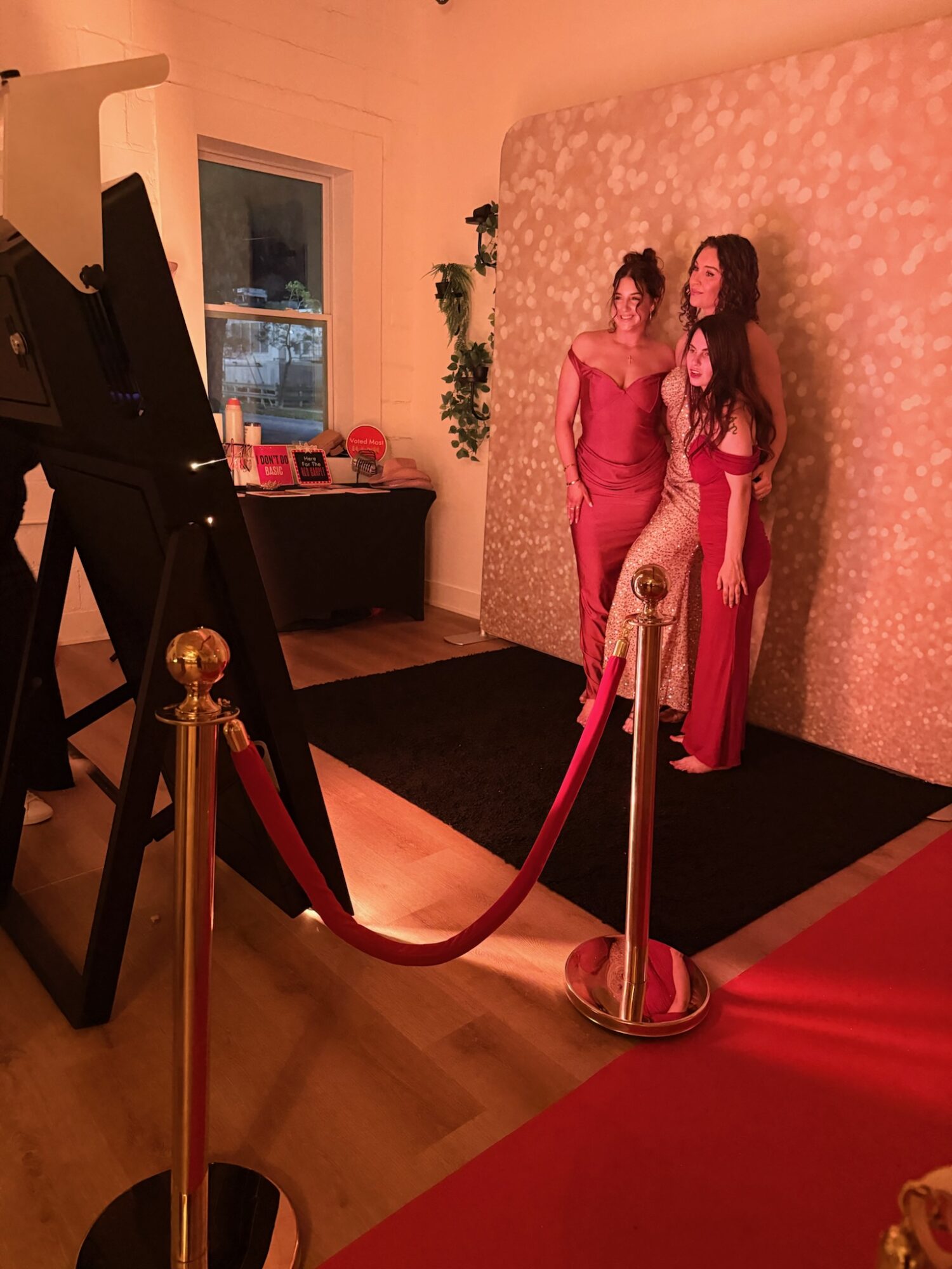 Two women in red dresses pose indoors near a pink floral backdrop, with a black carpet and velvet rope barrier.