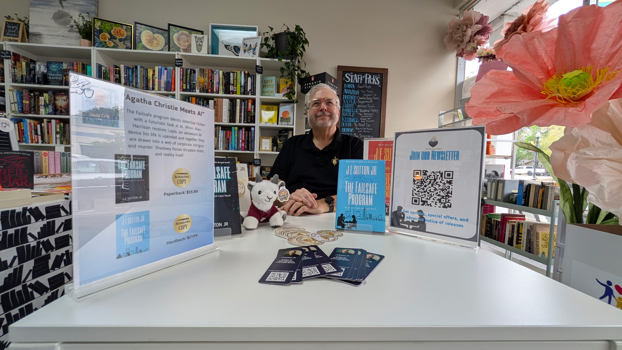 Person sitting at a table with books, posters, and a large pink flower in a library or bookstore setting.