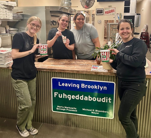 Four people standing behind a counter with a sign, holding food and a plant, smiling in a store.