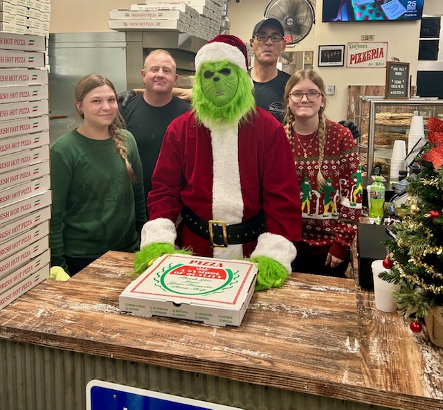 Person dressed as Santa with a green face and hands, standing behind a pizza box on a wooden counter, surrounded by four people.