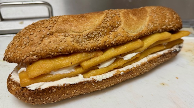 Sandwich with sesame seed bun, cheese, and white sauce on a white surface.