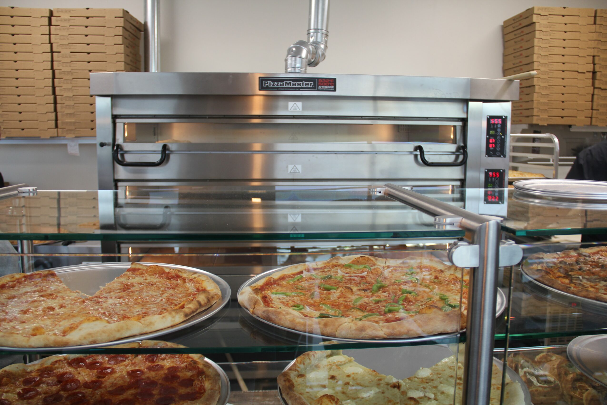 Commercial pizza oven with two pizzas on a glass display shelf in front, stacks of boxes in background.