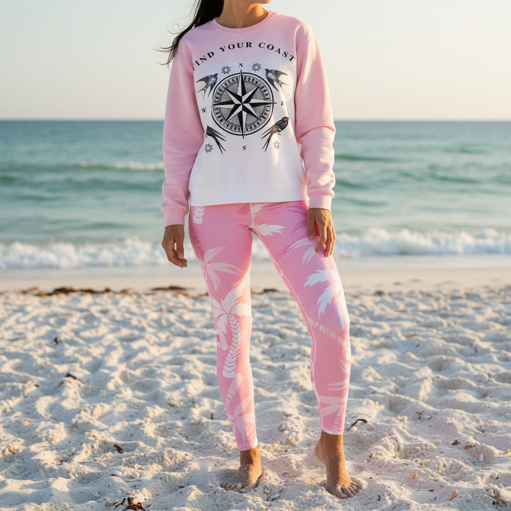 Person standing on sandy beach near ocean, wearing pink sweatshirt and matching leggings, facing camera.