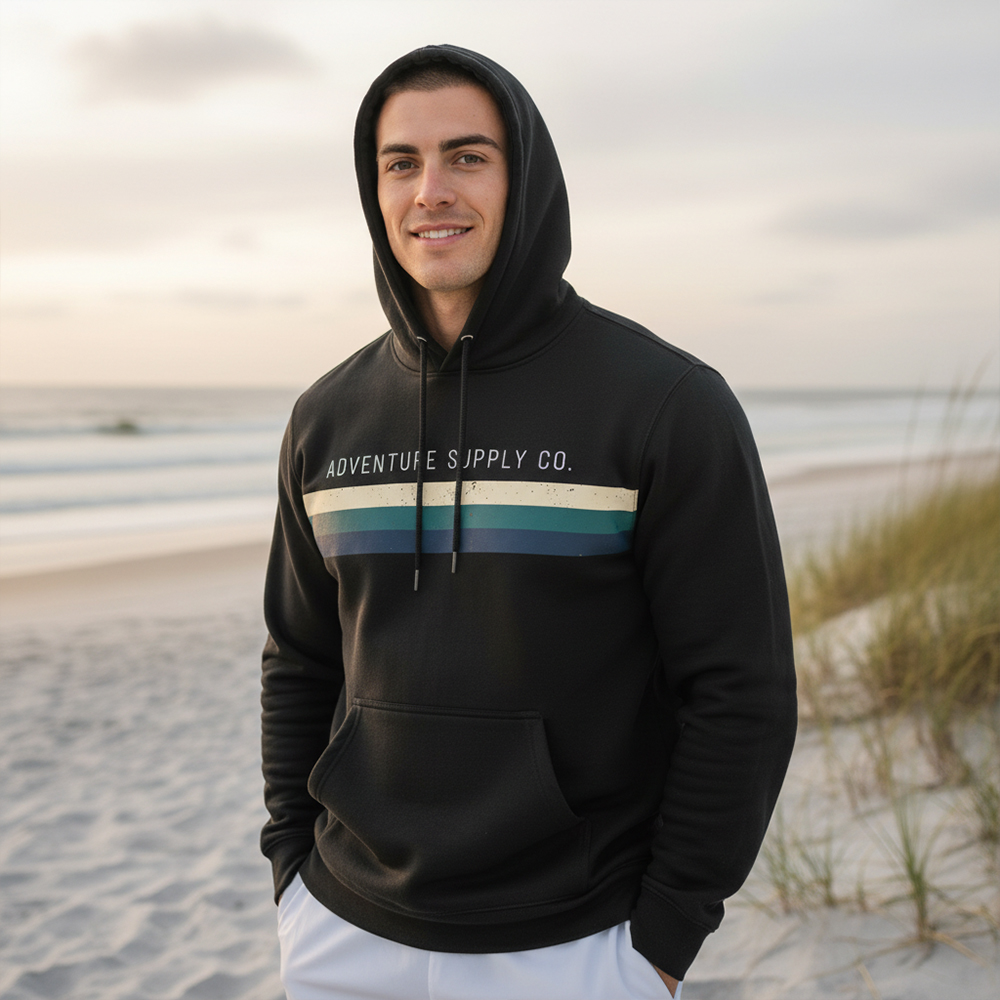 Young man in black hoodie standing on beach with sand and grass, smiling, overcast sky in background.