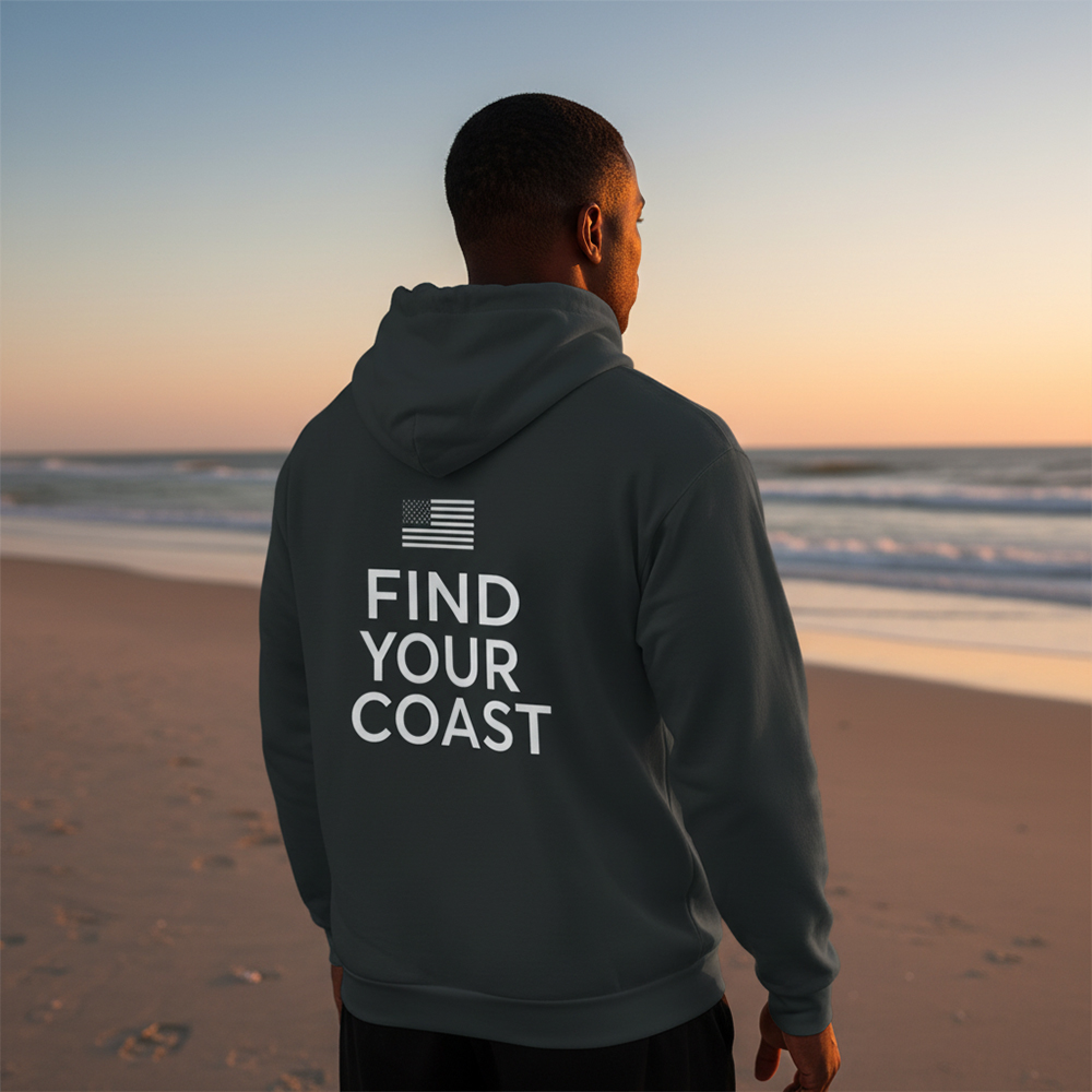 Person standing on beach at sunset, wearing hoodie with text and flag, facing ocean.