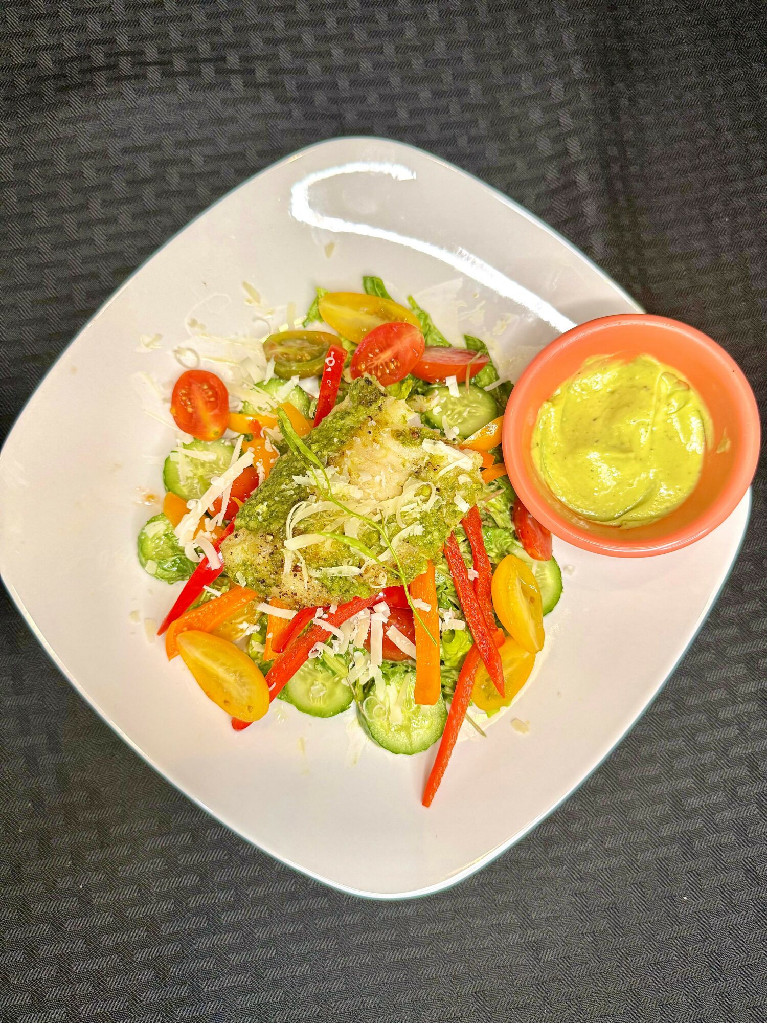 Mixed salad with cucumber, cherry tomatoes, red and yellow bell peppers, and a side of green dressing in a small bowl.