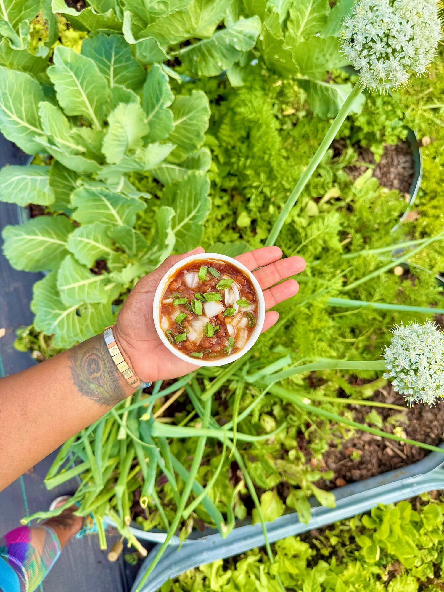 Person holding a bowl of salad with greens and vegetables in a garden with leafy plants and flowers.