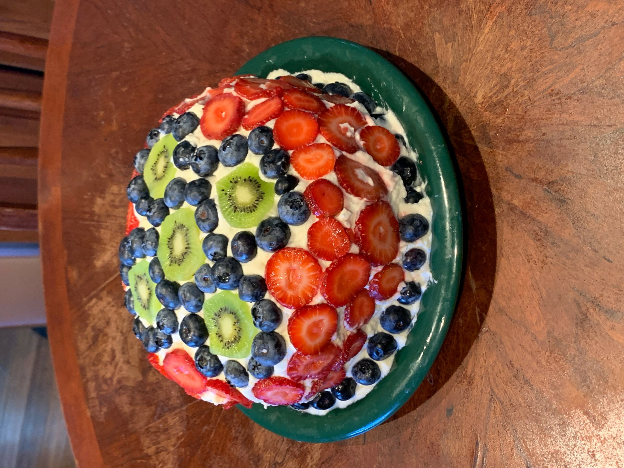 Round fruit cake topped with sliced strawberries, blueberries, and kiwi on a green plate, on a wooden table.