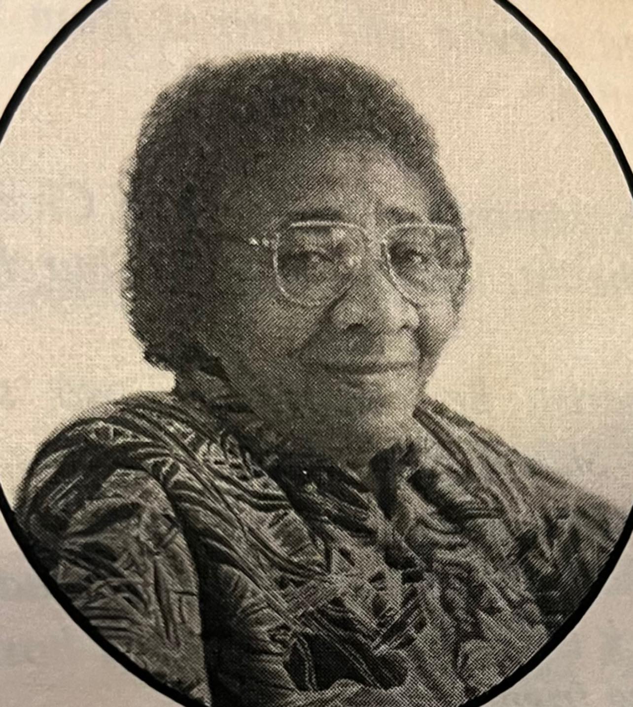 Older woman with glasses and short curly hair, wearing patterned clothing, smiling gently.