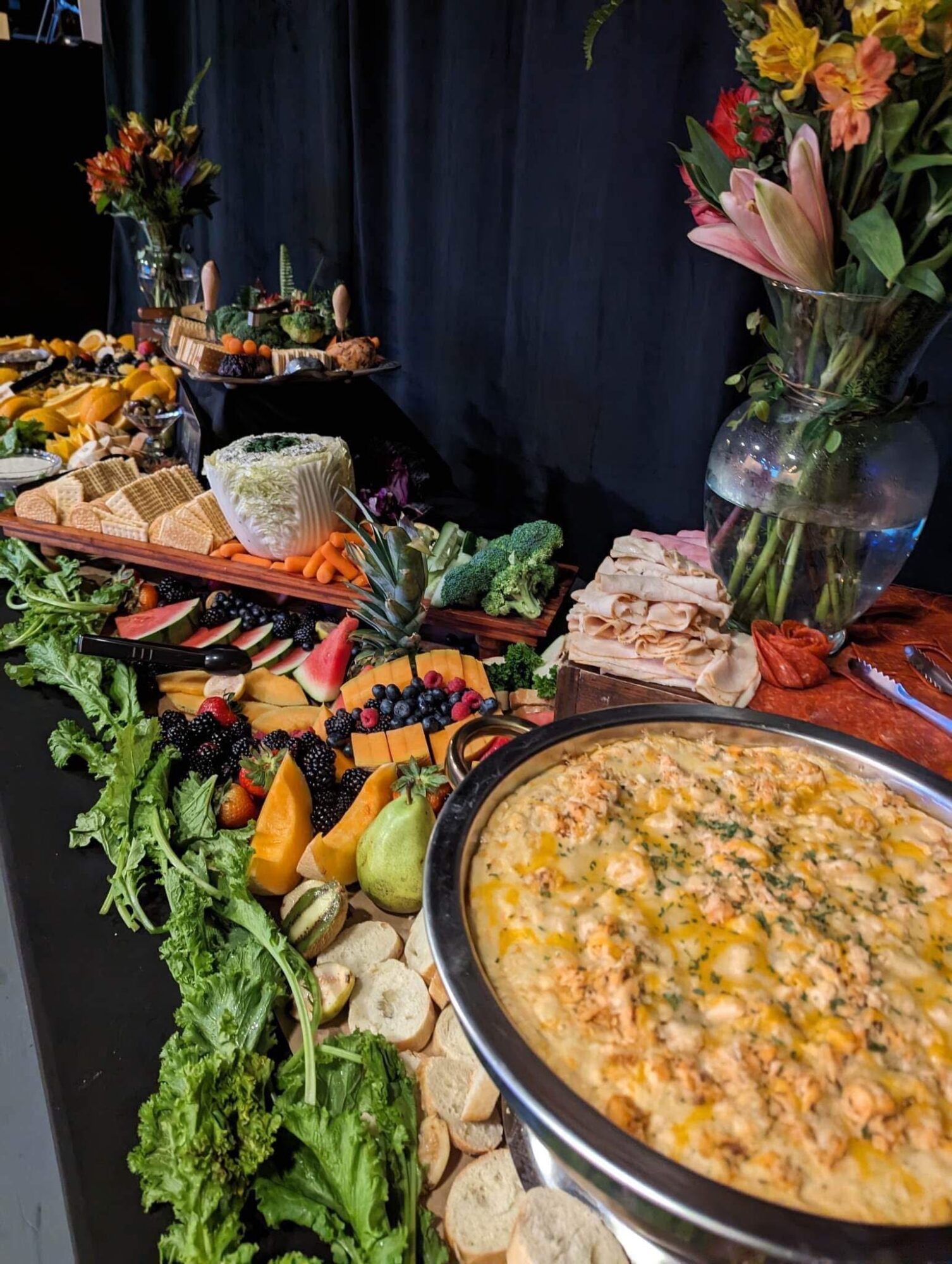 Buffet table with various dishes, fruits, and a large bowl of soup, decorated with flowers and greenery.