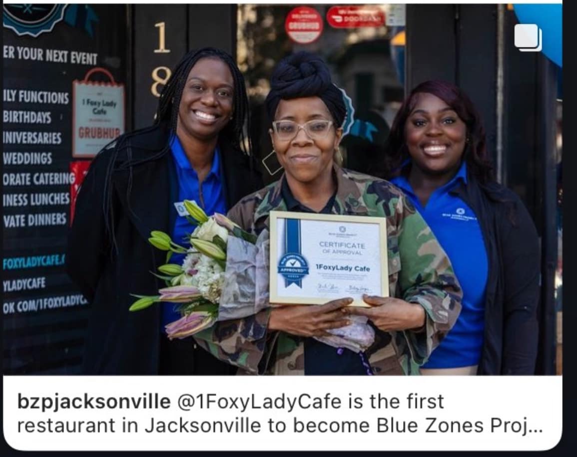 Three women standing outdoors, one holding a certificate and flowers, smiling at the camera.