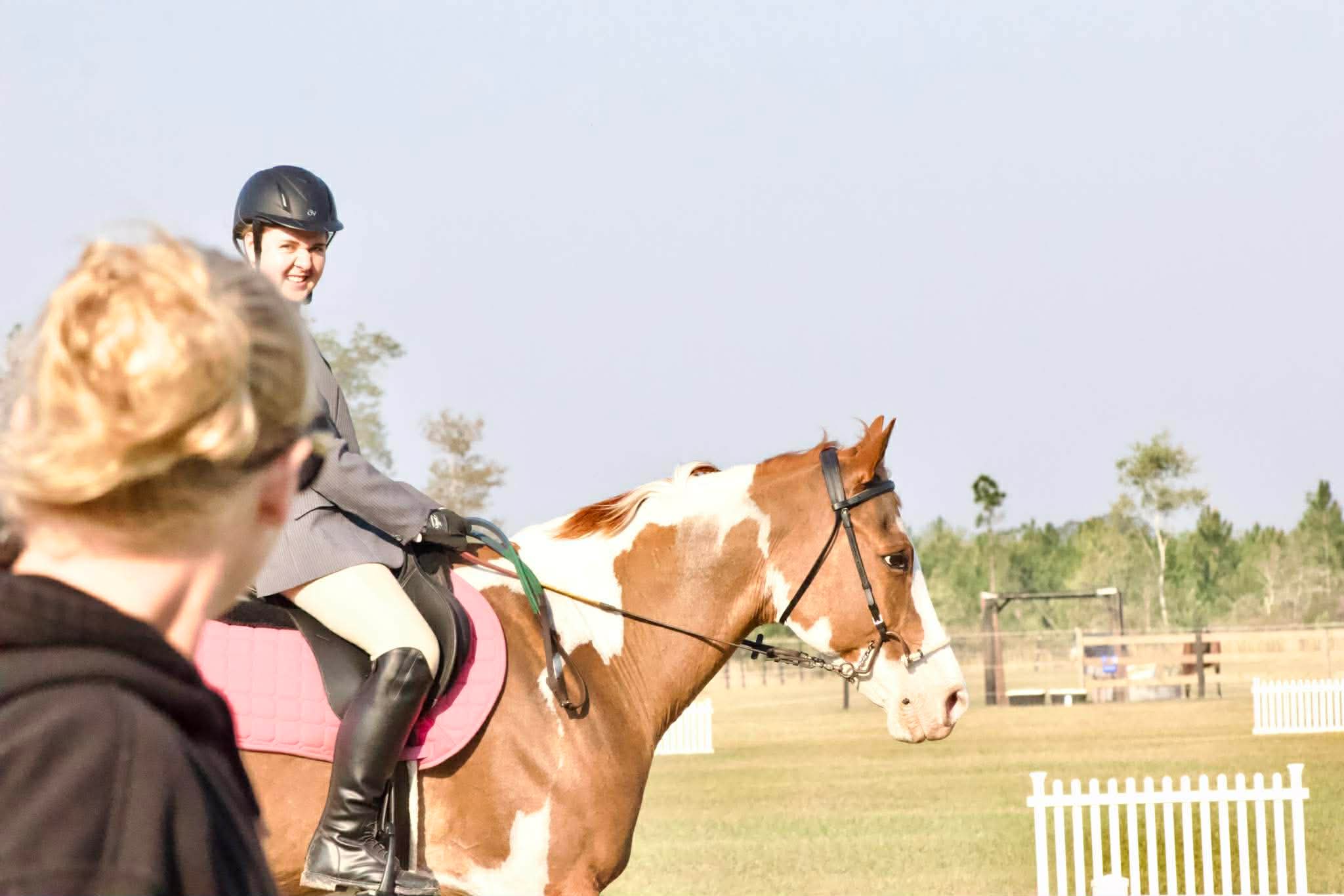 Person riding a horse with a helmet, another person with blonde hair in the foreground, outdoor setting with fencing and trees.