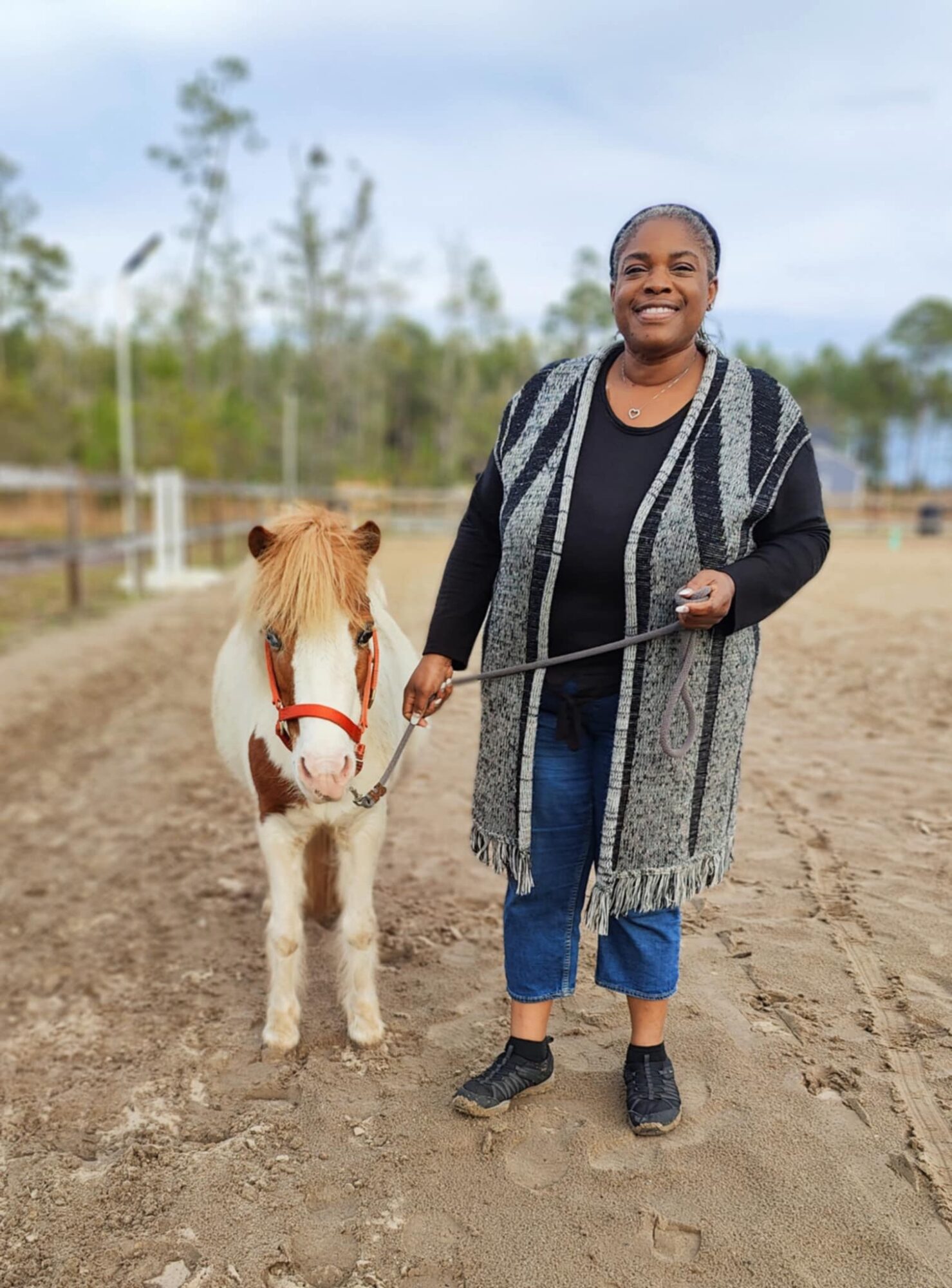 Person smiling holding a leash with a small horse on a dirt field outdoors.