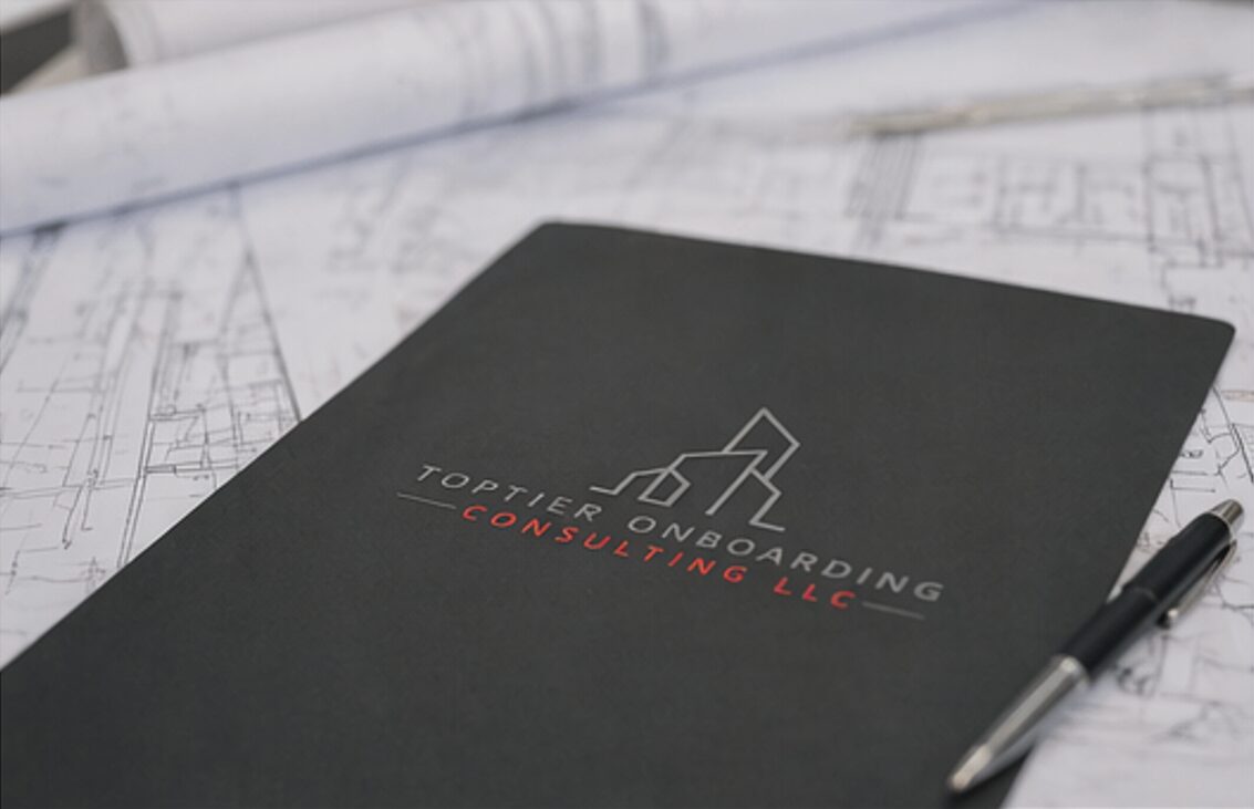 Black notebook with logo and text, pen, and architectural plans on table.