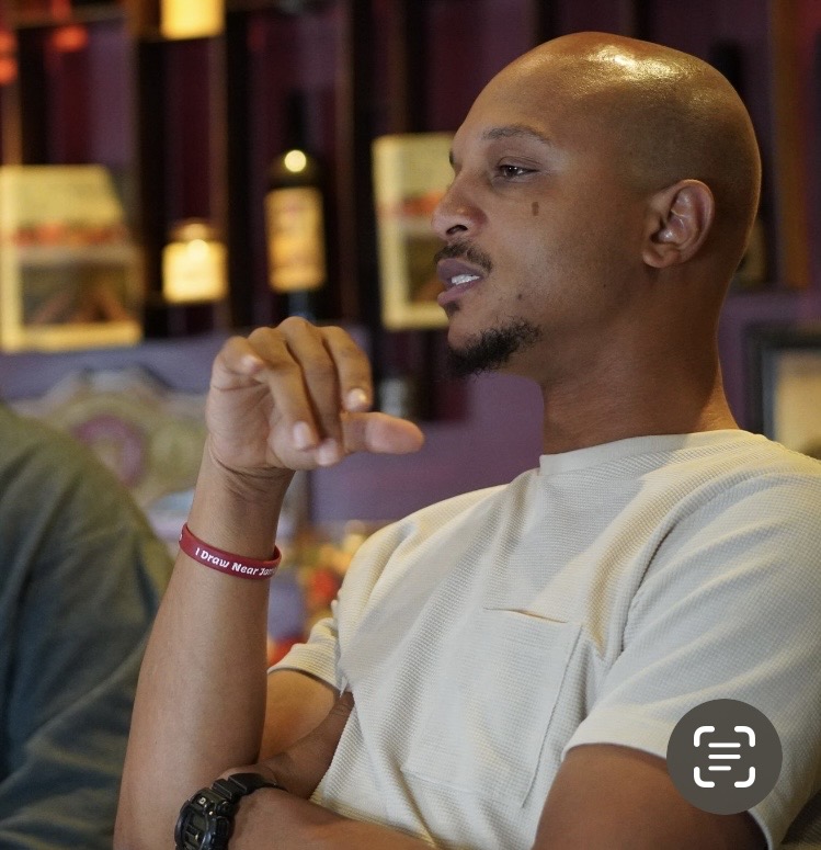 Man with a shaved head and goatee, wearing a white shirt, sitting indoors with shelves in the background.