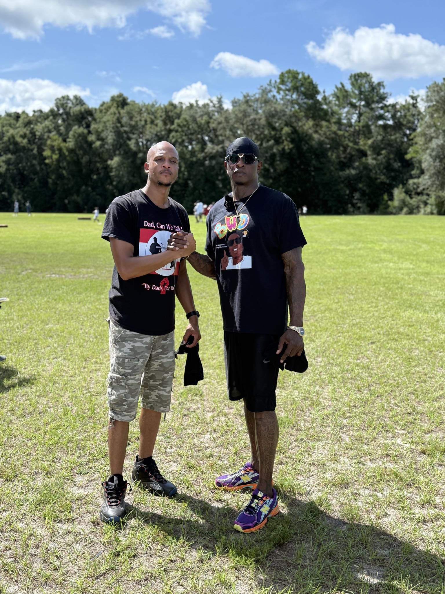 Two men standing outdoors on grass with trees and blue sky in background, holding drinks and posing for photo.